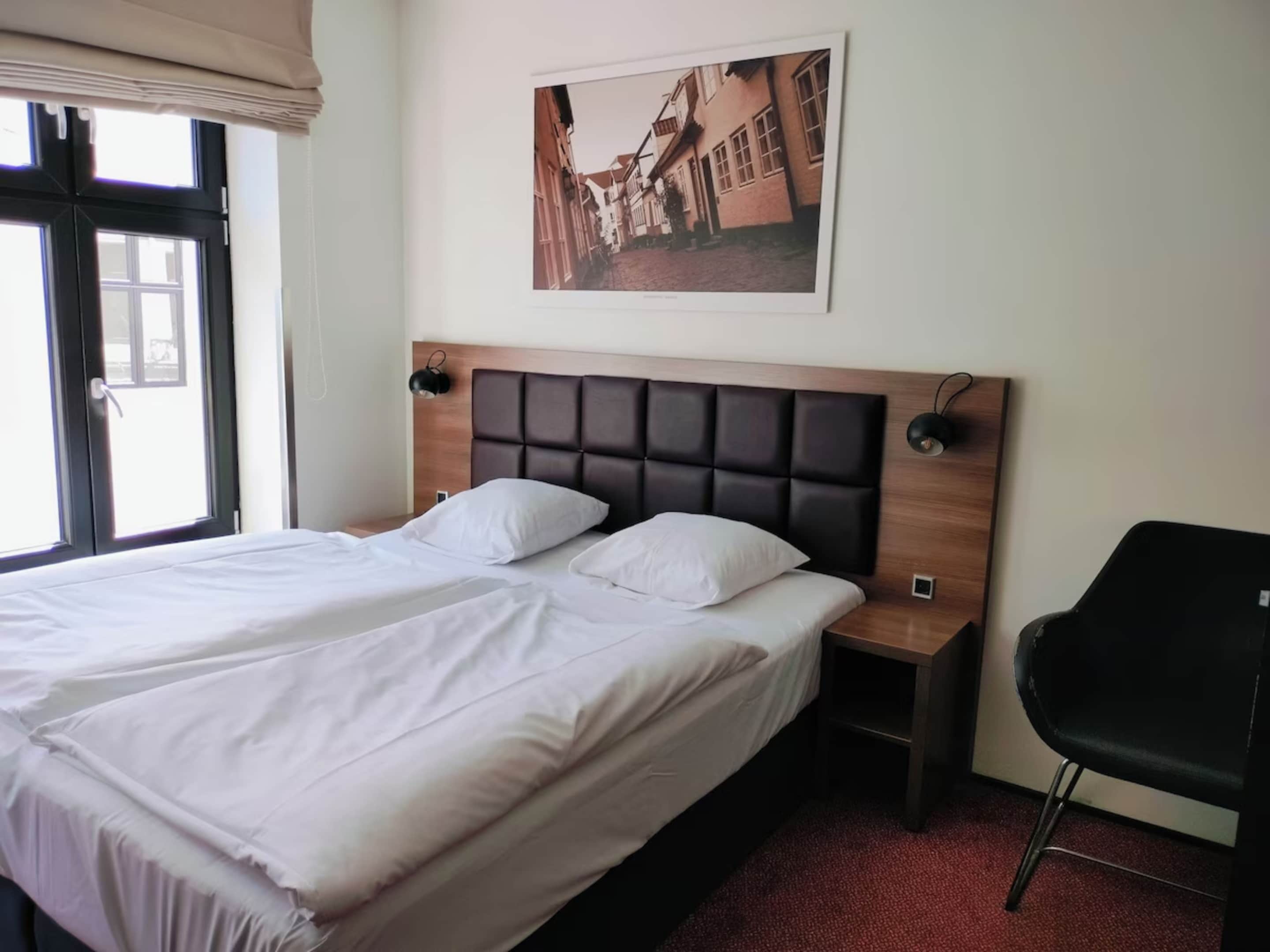 Una habitación de hotel con una cama grande, cabecera, lámparas de noche, una silla y un cuadro en la pared.