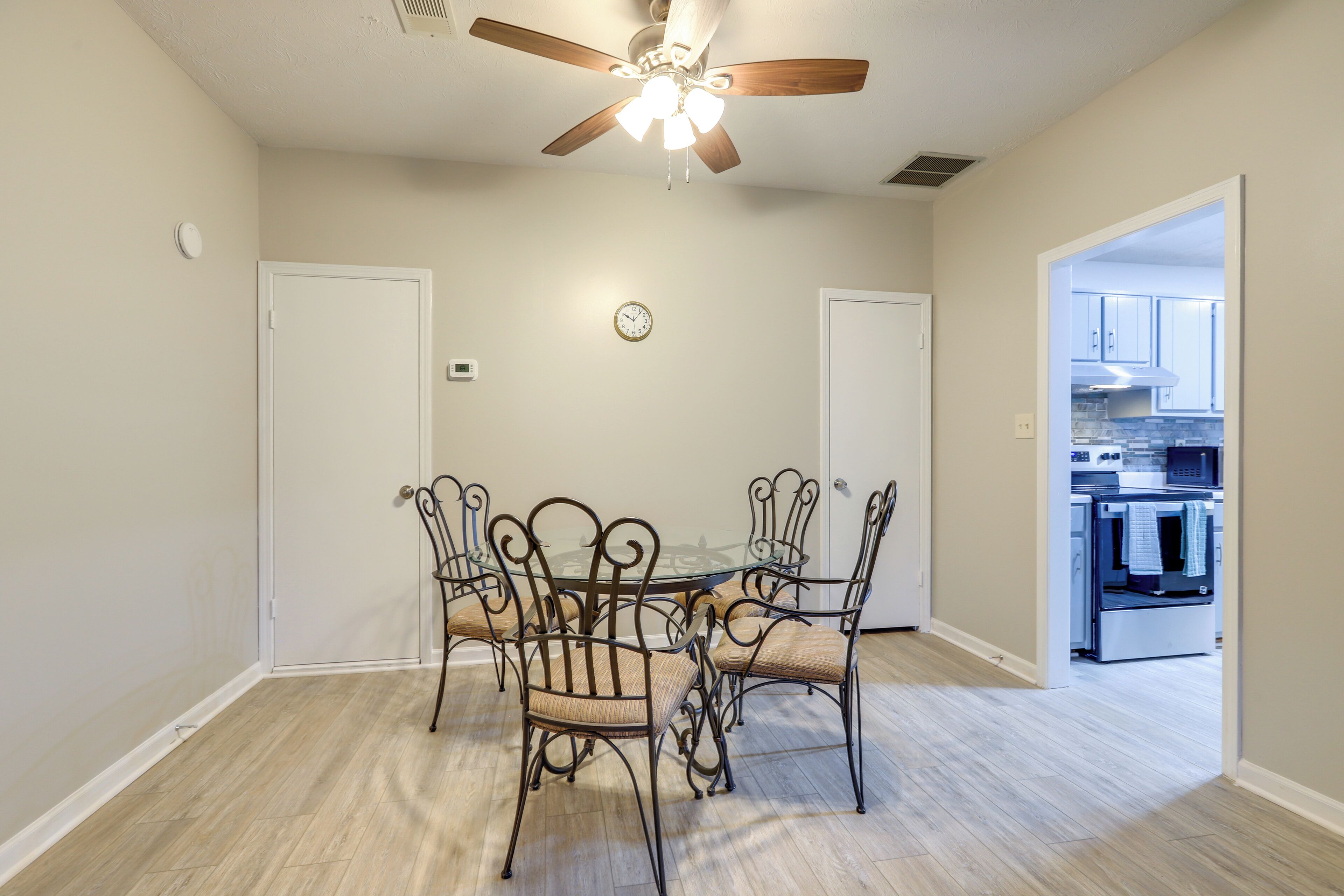 A dining area with a round table and four chairs, a ceiling fan, a clock, and an adjacent kitchen.