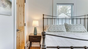 A bedroom with a wooden door, a bedside table with a lamp, a bed with patterned bedding, and a window with blinds. - Walk to Harbor, Eats & Shops! Greenport Retreat (Greenport)