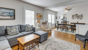 A modern living room with a gray sofa, ottoman, and coffee table. There is a dining area with a table and chairs, and a ceiling fan. - Walk to Harbor, Eats & Shops! Greenport Retreat (Greenport)