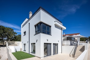 Ein modernes, zweistöckiges Haus mit flachem Dach, großen Fenstern und einer Treppe, die zu einer oberen Ebene führt.