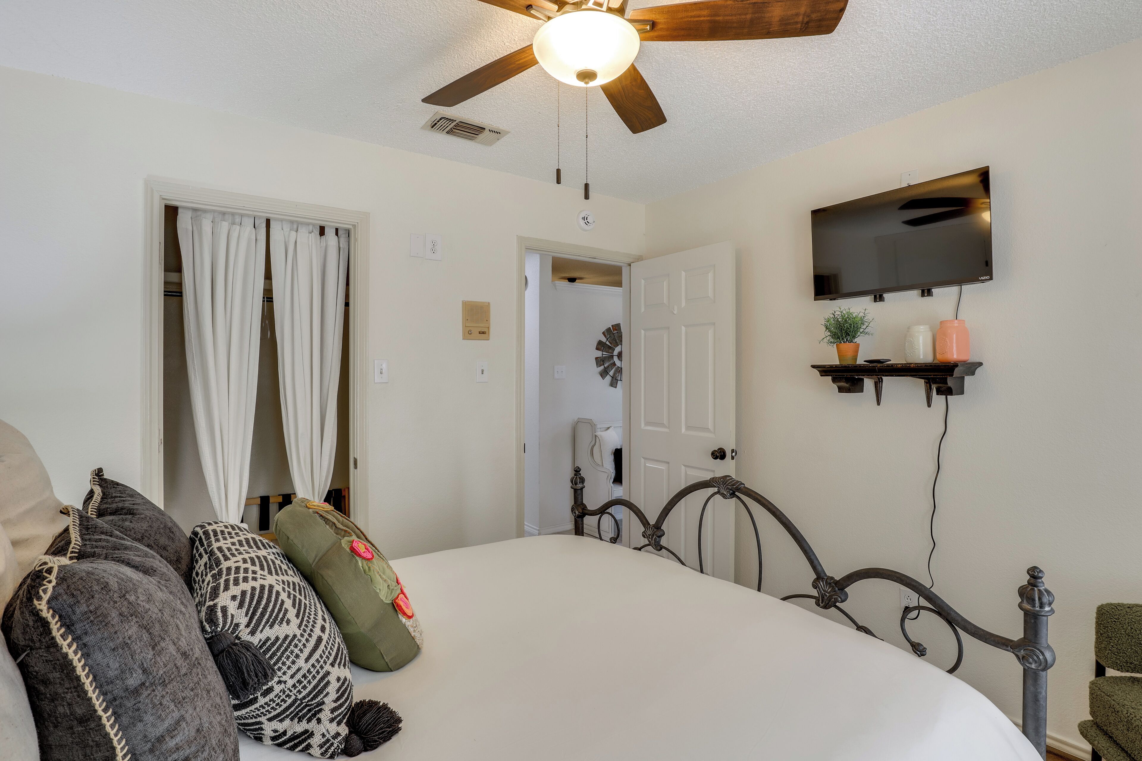 A bedroom with a bed, a ceiling fan, a TV mounted on the wall, and a door leading to another room.
