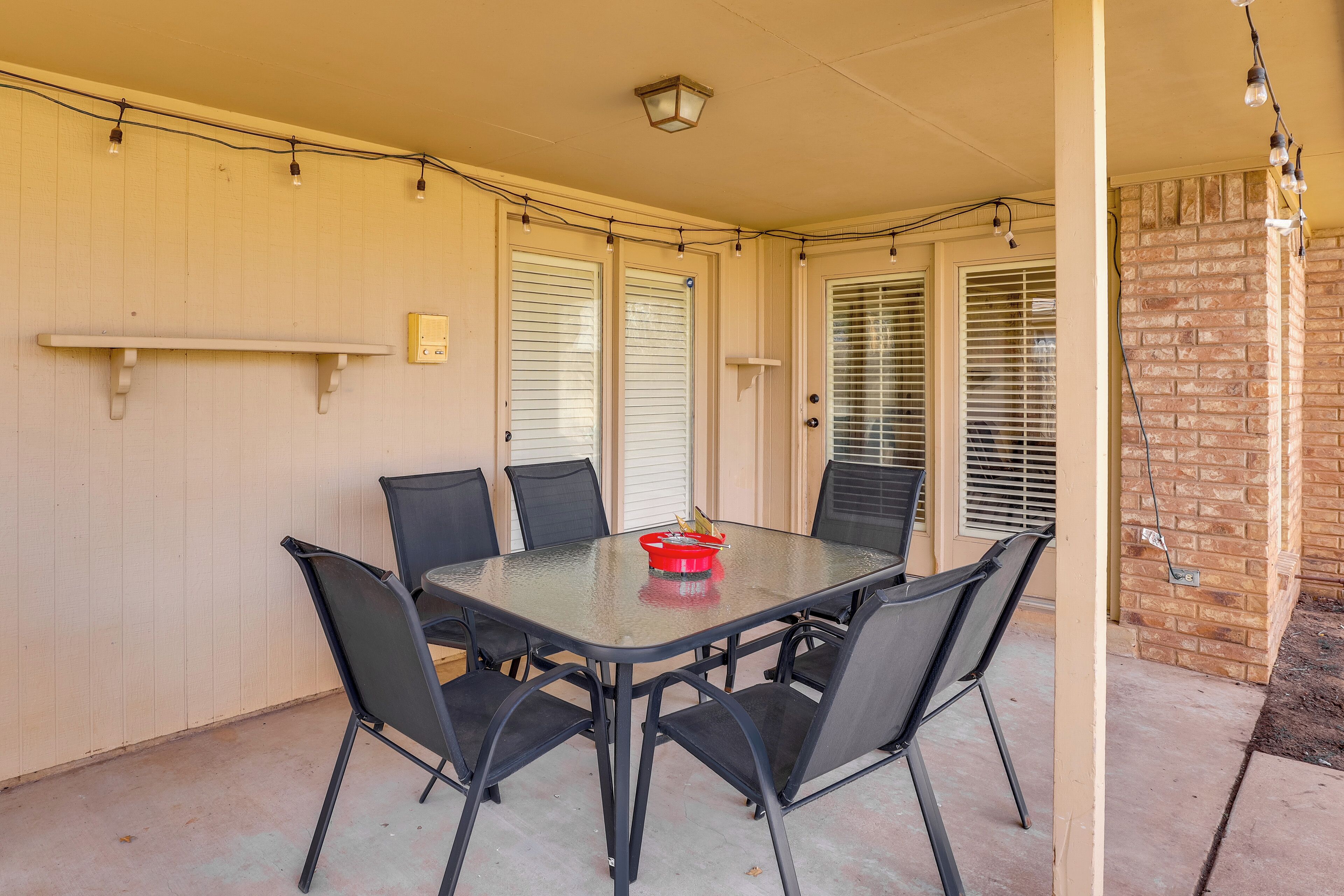 Un patio techado con una mesa de vidrio y seis sillas, un cuenco pequeño rojo y luces colgantes.