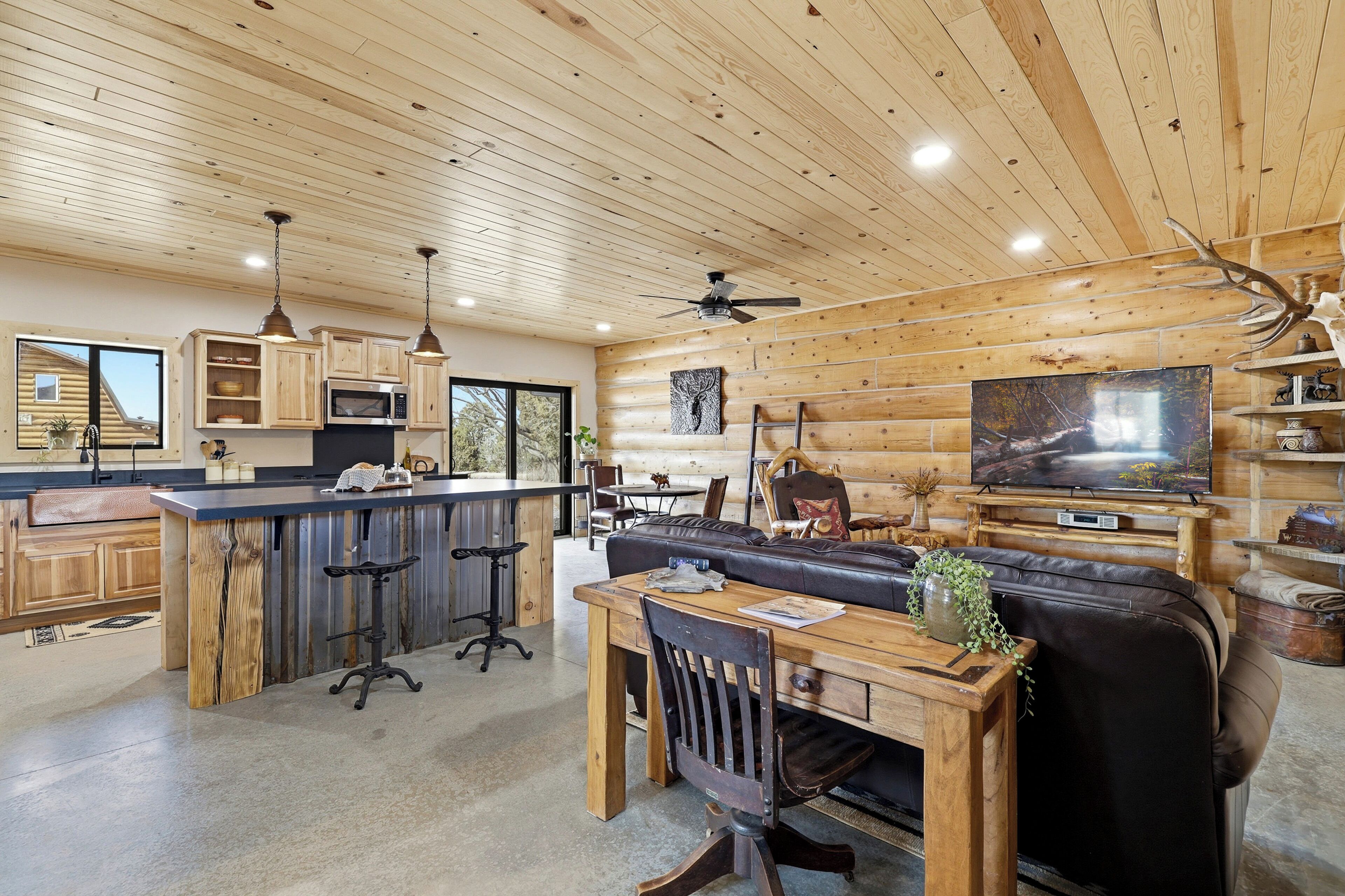 A rustic cabin interior with a wooden ceiling, a dining area with a wooden table and chairs, a bar area, a living room with a sofa and a television, and a kitchen with wooden cabinets and a microwave.
