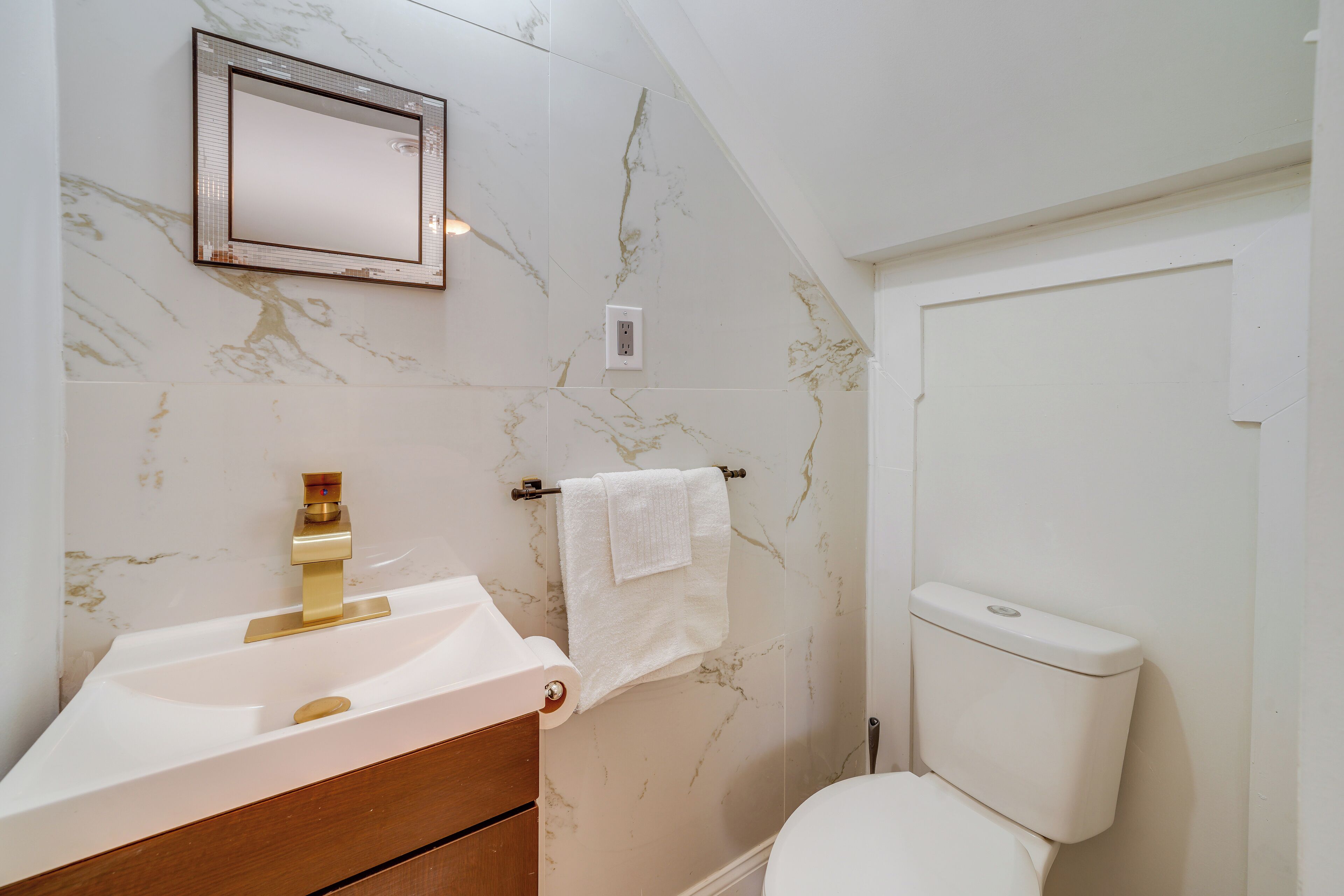 Un bany amb una paret de marbre, un lavabo amb un aireix d'or, un vàter blanc i una tovallola penjada en un suport de tovalloles.