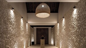 Een hal met structuurgevoegde stenen muren, een centrale kolom met een decoratief object, en een grote geweven lamp.