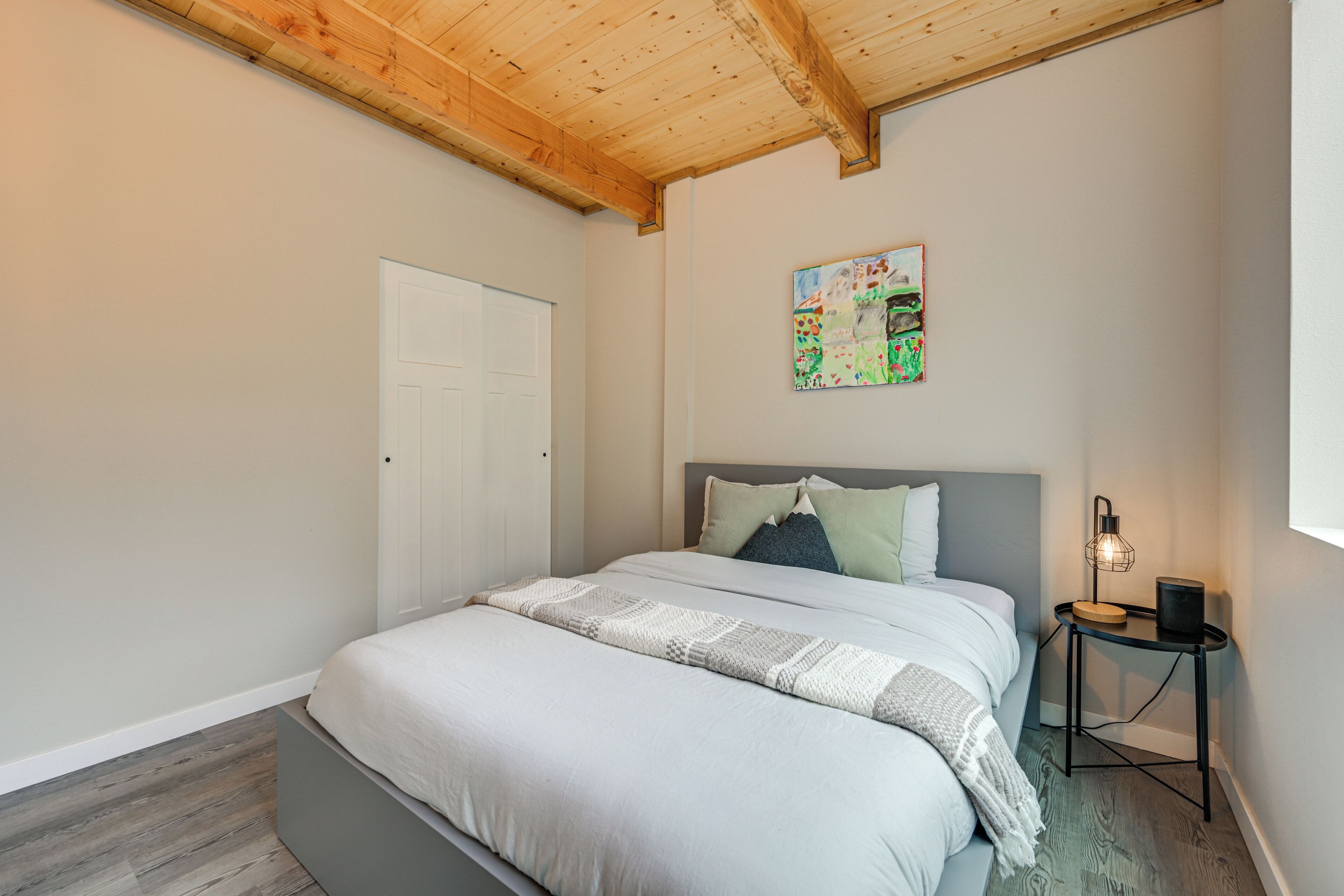 A bedroom with a bed, a bedside table with a lamp, a door, and a painting on the wall.
