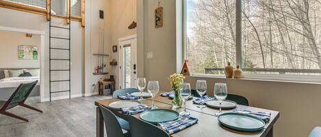 Un comedor con una mesa de madera dispuesta para cuatro, sillas azules y vistas a árboles a través de un ventanal grande.