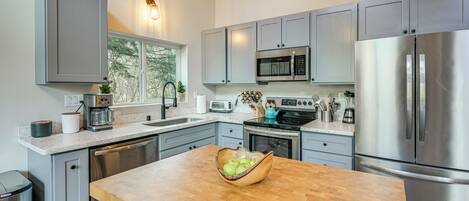 A modern kitchen with grey cabinets, stainless steel appliances, a wooden island, and a window with a view of trees.