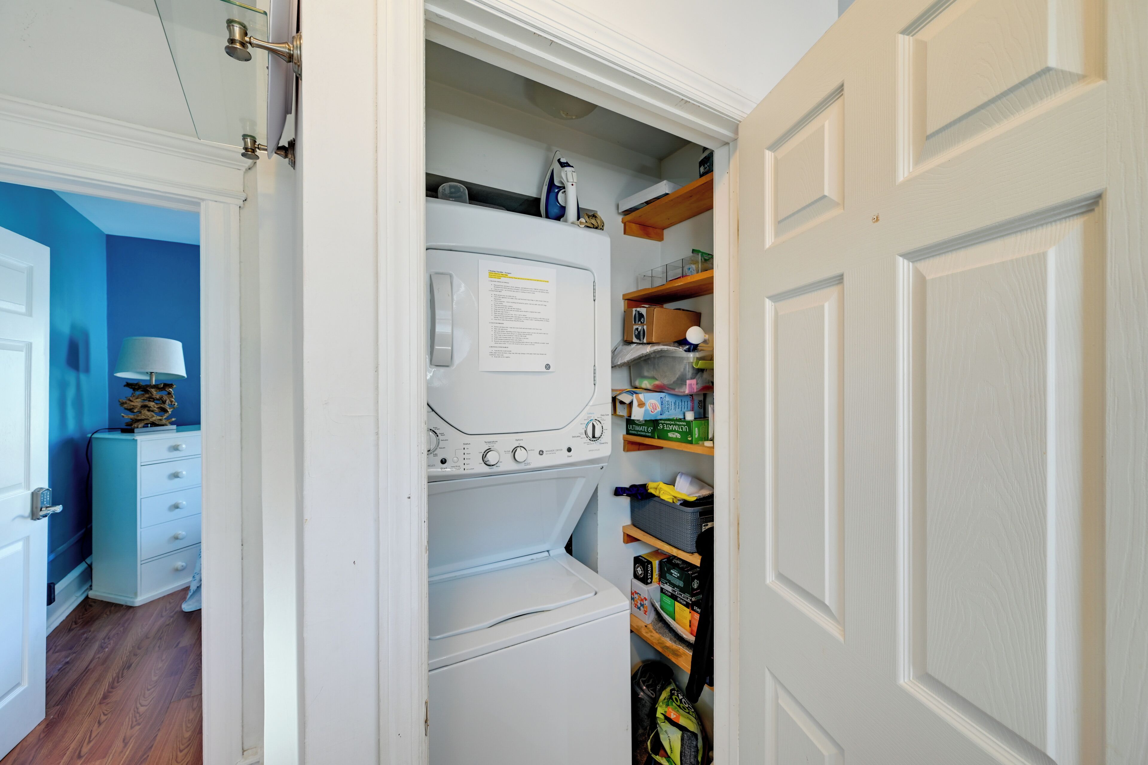 A laundry room with a front-loading washer and dryer, shelves with cleaning supplies, and a door leading to another room.