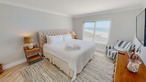 A bedroom with a large bed, a bedside table with a lamp, a chair, a TV, and a window with blinds. - Destin On The Gulf 506 2 Bedroom Condo (Destin)