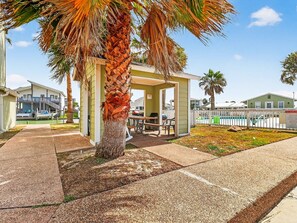 Et overdækket udendørs opholdsområde med et bord, stole, en palme og en swimmingpool i baggrunden.