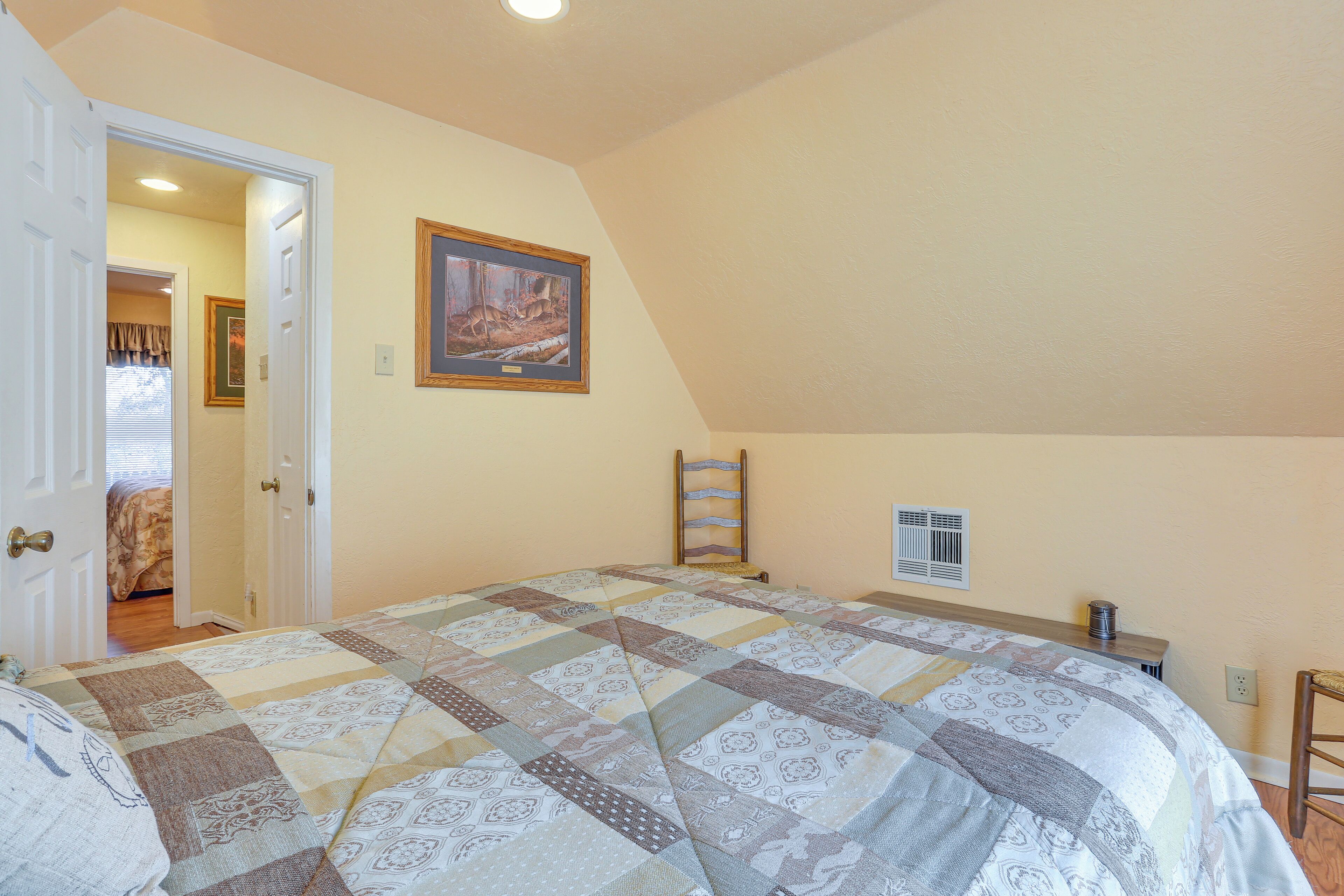 A bedroom with a large bed, a door leading to another room, a framed picture on the wall, and a small table with a lamp.