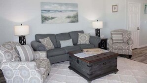 Un salon avec un canapé gris, des fauteuils à motifs, une table basse en bois et un tableau sur le thème de la plage accroché au mur.