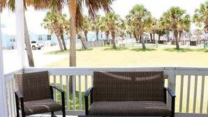 Une terrasse en bois avec deux fauteuils en osier et un banc, donnant sur une pelouse agrémentée de palmiers et sur laquelle on aperçoit un bâtiment au loin.