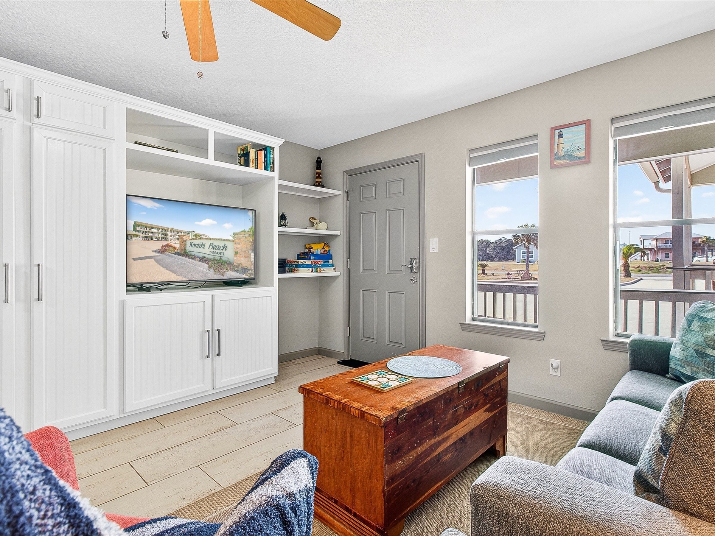 A living room with a television, white cabinets, a wooden coffee table, and a view of a beach through a window.