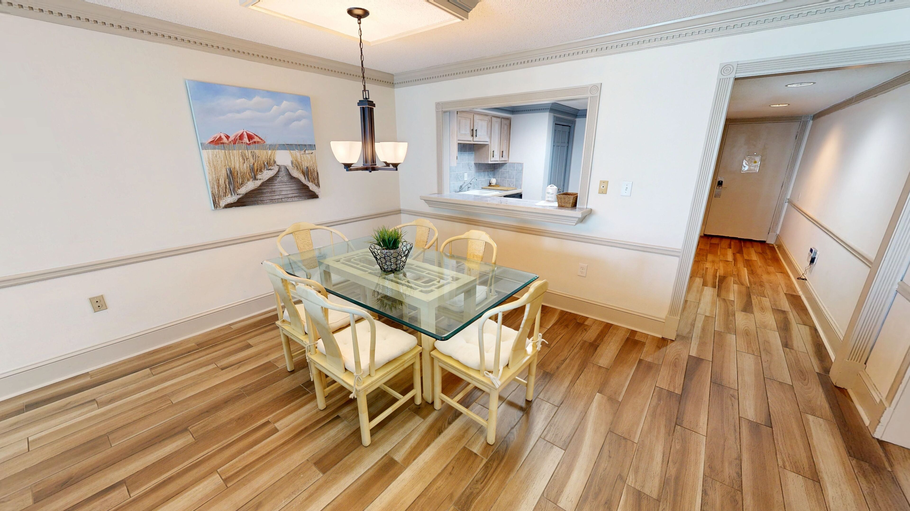 A dining area with a glass table and chairs, a painting on the wall, and a kitchen visible through an open doorway.
