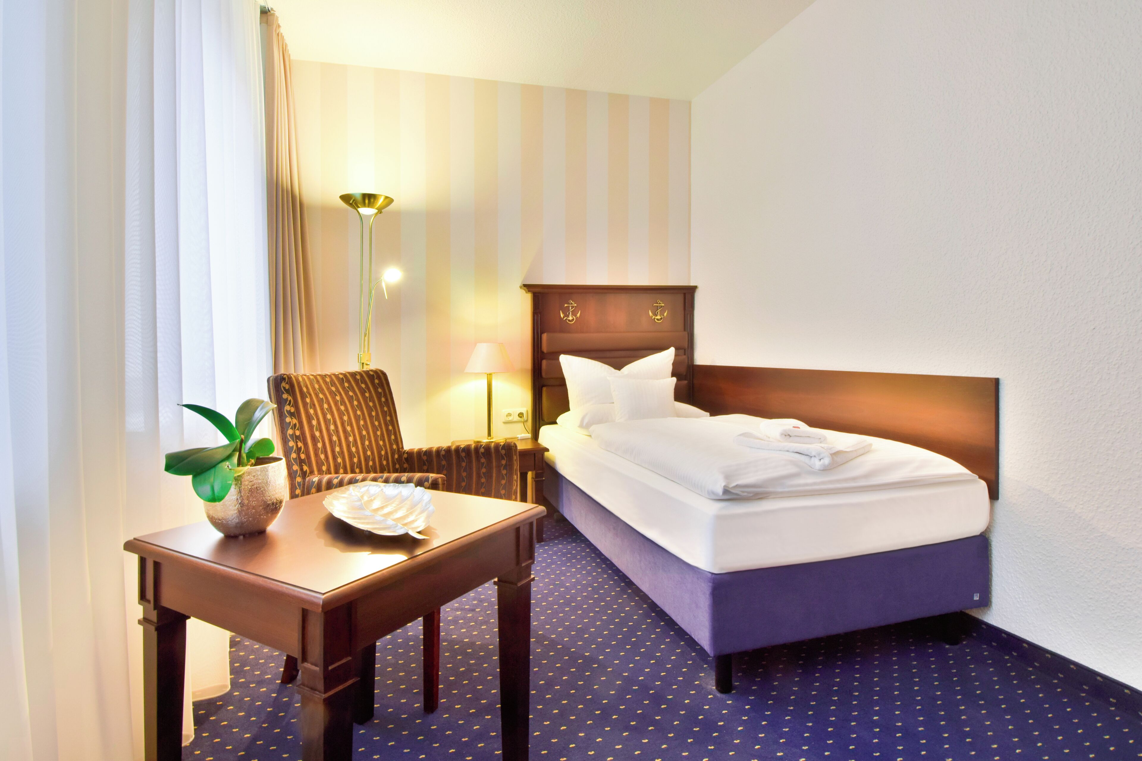 A hotel room with a bed, a desk, a chair, a lamp, and a potted plant.