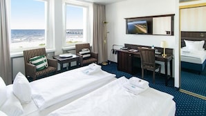 A hotel room with two beds, a desk, a chair, a TV, and a view of the ocean. - Promenandenhotel Admiral (Seebad Bansin)