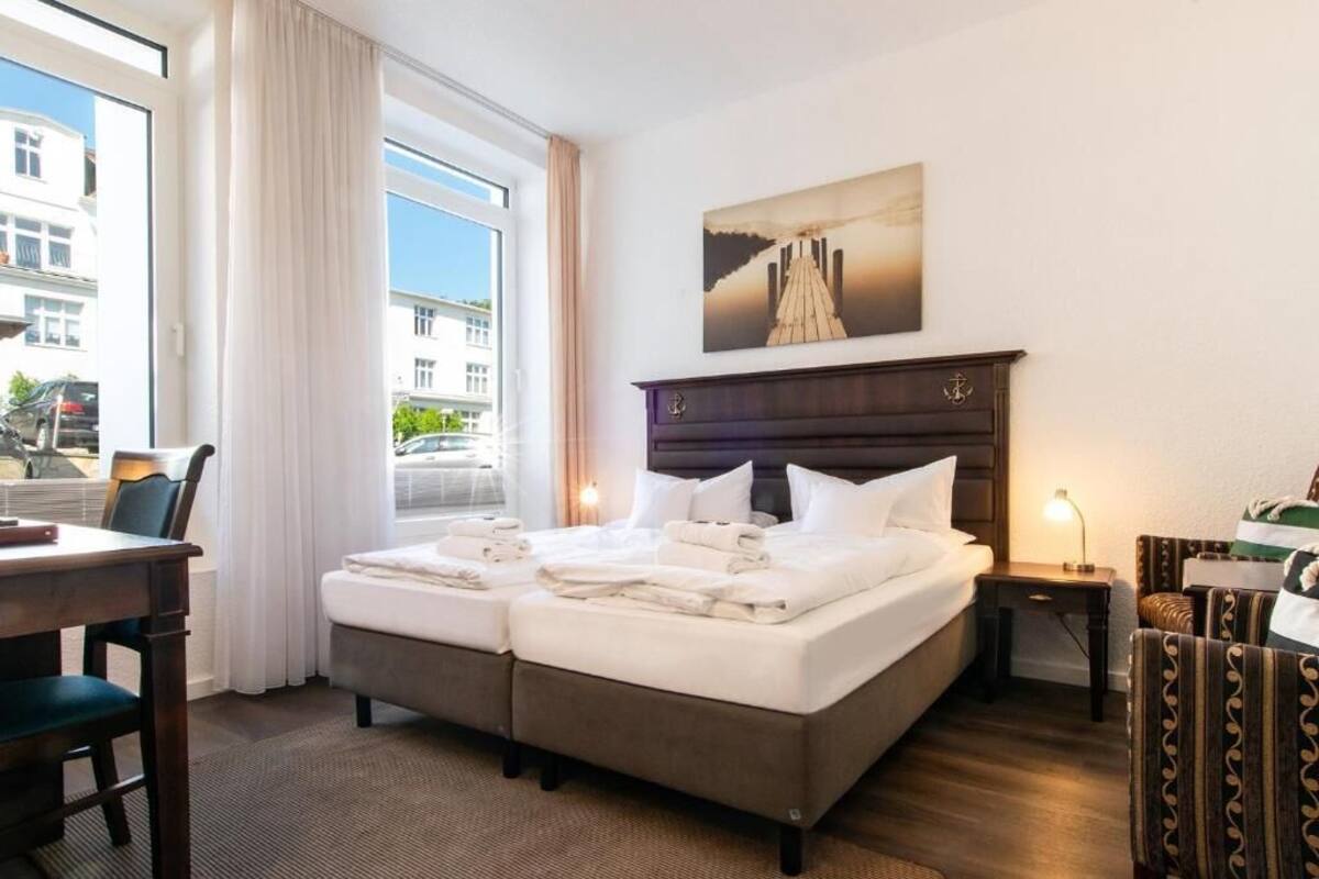 Une chambre d’hôtel avec un grand lit, un bureau et une chaise. Il y a une fenêtre avec des rideaux, un tableau au mur et une lampe sur la table de chevet.