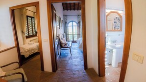 A narrow hallway with a bed, a chair, and a bathroom visible through an open door. - Pero – Two Level Apartment in Tuscan Farmhouse with Swimming Pool, 2 Bedrooms (Castelfiorentino)