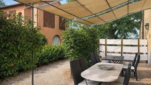 En udendørs terrasse med et bord og stole, en pergola der giver skygge, og en bygning med et gittervindue i baggrunden.