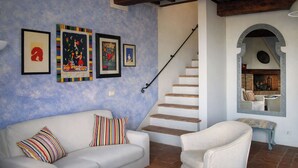 A living room with a white sofa, a striped pillow, and a staircase leading to a dining area with a brick fireplace. - Plum – Two-Level Apartment in Tuscan Farmhouse with Swimming Pool, 2 Bedrooms (Castelfiorentino)