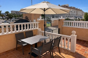 A terrace with a table and chairs, a beige umbrella, and a view of buildings in the background. - Stylish South House in Villamartin - Solarium and Poolview Terrace (Orihuela)