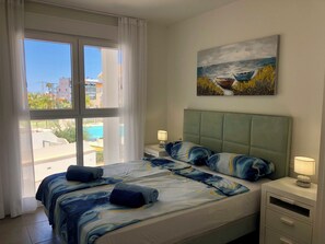 Ett hotellrum med en stor säng, en målning av båtar, utsikt över byggnader och en pool utanför fönstret.