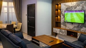 Een moderne woonkamer met een grote tv aan de muur, een bank, een salontafel en een eethoek met een tafel en stoelen.