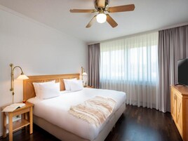 Habitación de hotel con una cama grande, cabecera de madera, dos lámparas de noche, ventilador de techo, televisor sobre un mueble de madera y una ventana con cortinas.