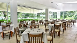 A spacious dining area with multiple tables and chairs, large windows offering a view of greenery outside. - VILLAS HOLIDAYS (Kiten)