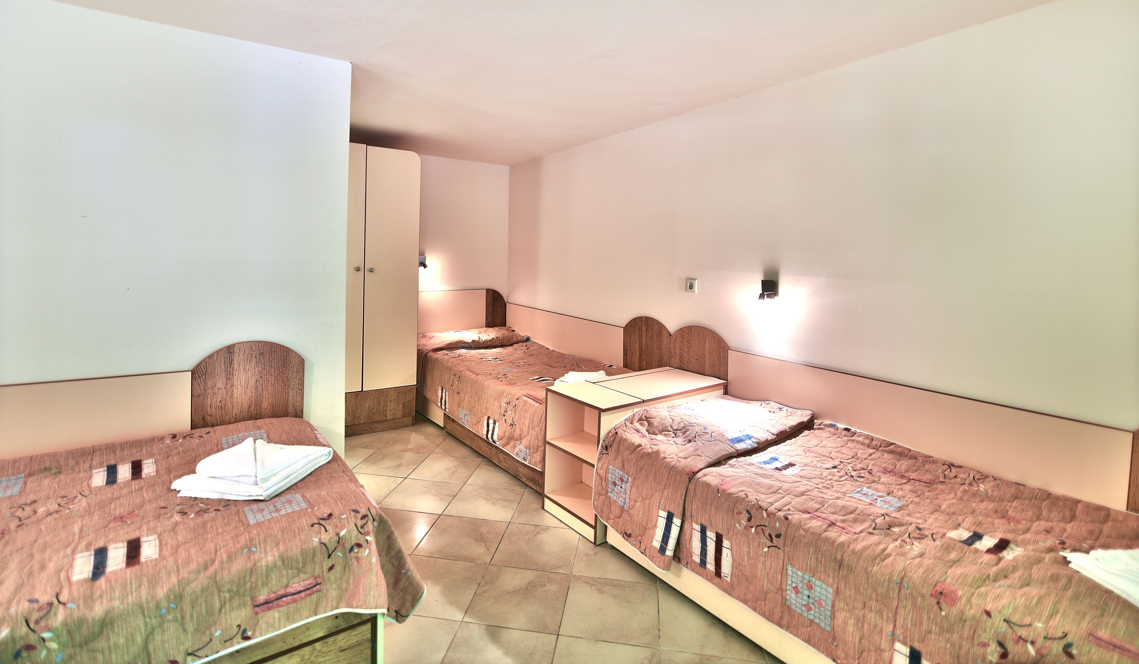 A dormitory room with four single beds, each with a wooden headboard and a quilted bedspread. There is a wardrobe with two doors and a small table with a chair.