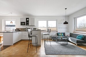 Una cocina moderna con armarios blancos, una zona de comedor con taburetes de bar, un sofá gris y una mesa de centro redonda.