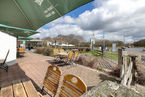 A patio area with wooden benches and tables, a covered seating area with umbrellas, and a view of a road and trees in the background.