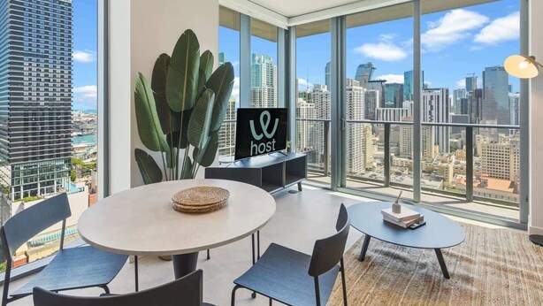 A modern hotel room with large windows, a round dining table, and a sign reading 'host'. - Chic 2BR | Pool | Free Parking | Miami Worldcenter (Miami)