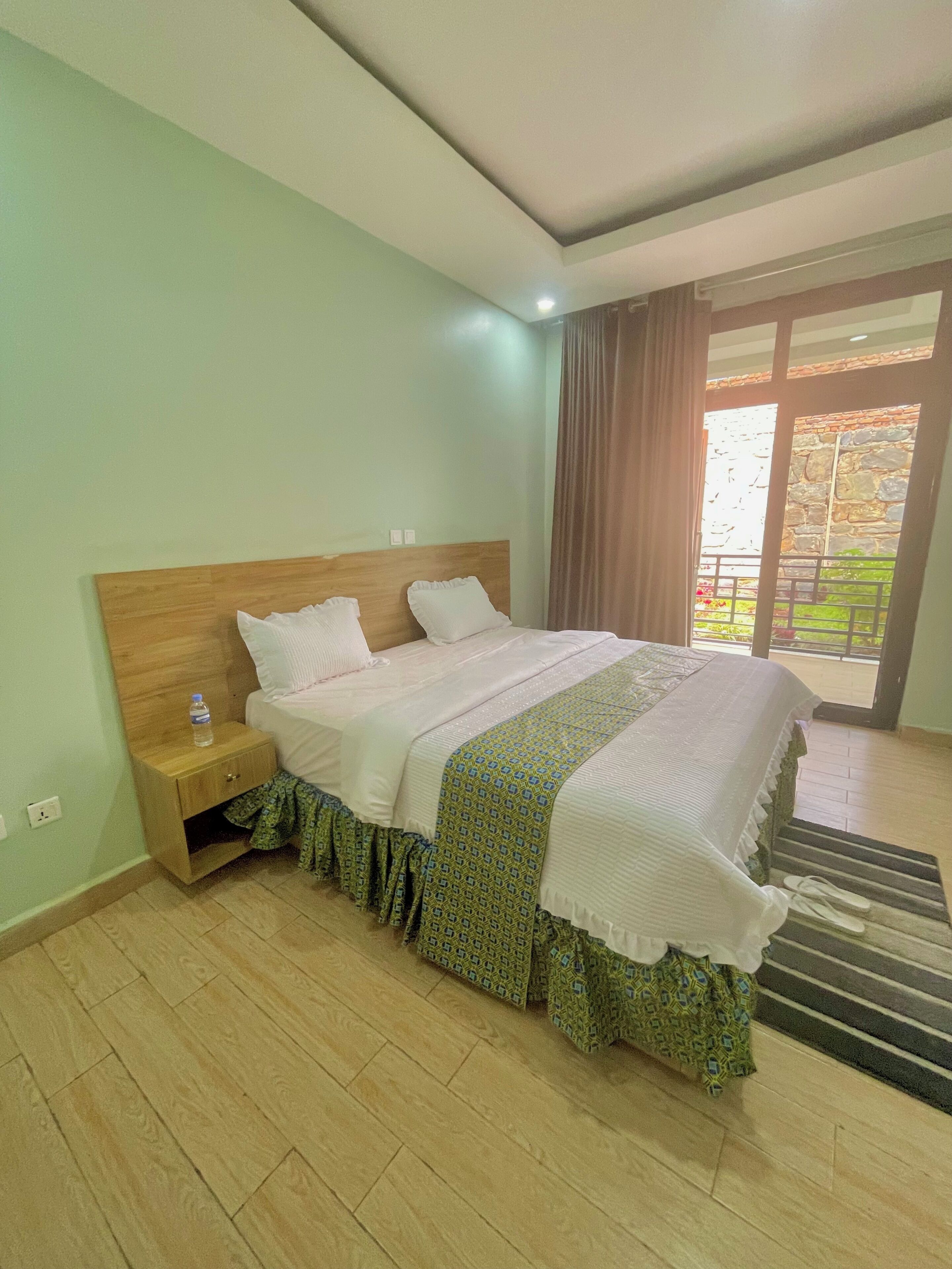 Habitación de hotel con cabecera de madera, cama con colcha estampada, mesita de noche con una botella de agua y vistas a un jardín a través de una puerta de vidrio.