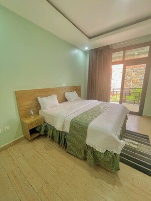 A hotel room with a wooden headboard, a bed with a patterned bedspread, a nightstand with a water bottle, and a view of a garden through a glass door. - Ibigabiro Hotel (Kagano)