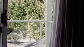 A room with a white chair near a window, curtains partially drawn, and a view of trees and a building outside.