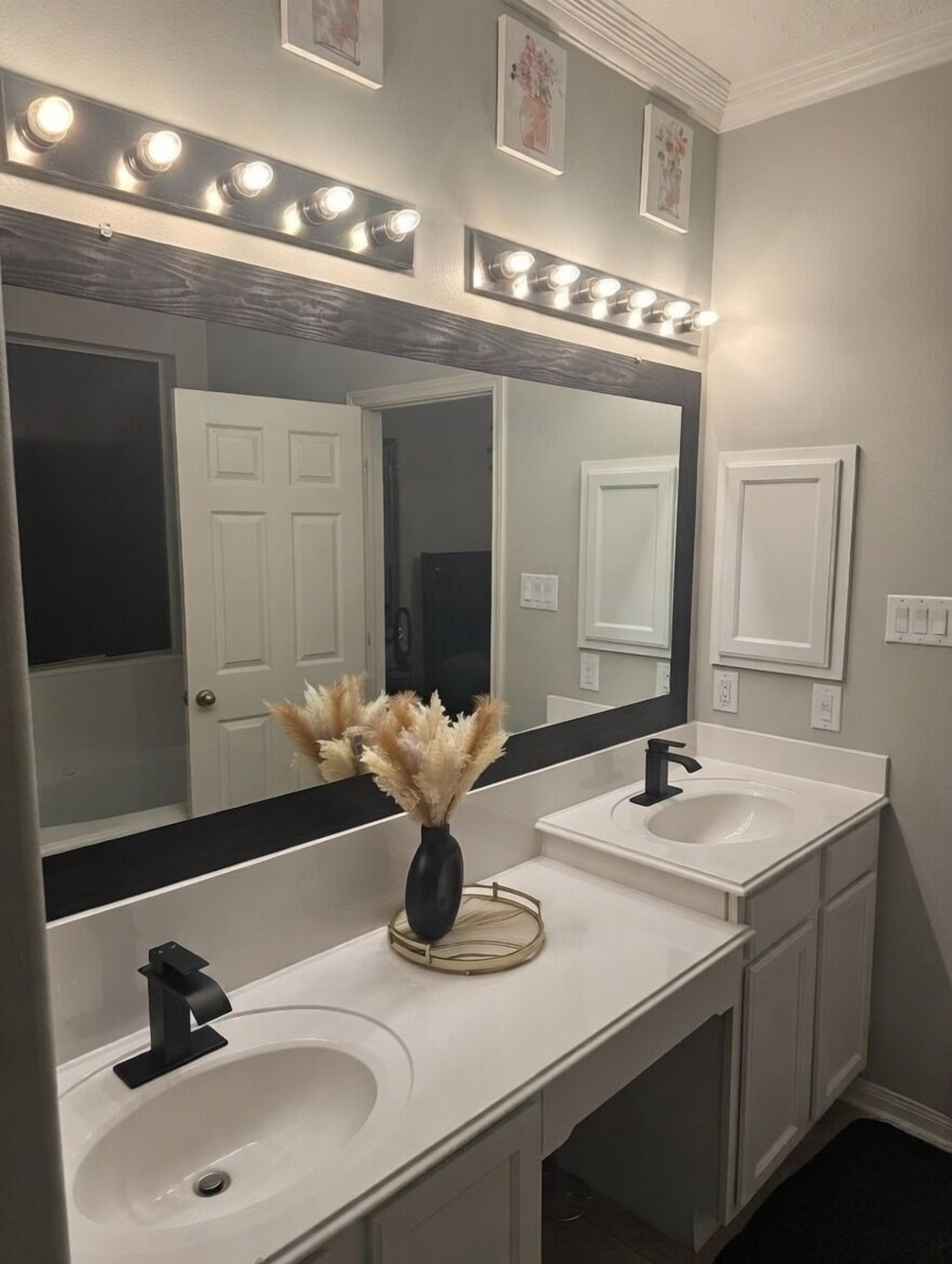 A bathroom with two sinks, a large mirror with lights, and a vase with dried flowers.