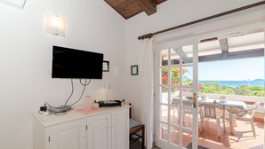A room with a TV mounted on the wall, a cabinet with various items, a chair, and a view of the outdoors through a sliding glass door. - Apartment 'Trilocale Taverna 80m from Beach' with Sea View, Private Terrace & AC (Porto Taverna)