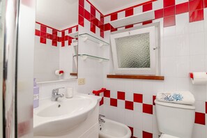 Ein Badezimmer mit weißem Waschbecken, einem Bidet, einer Toilette mit dekorativem Deckel und einem Spiegel mit strukturierter Glasplatte.