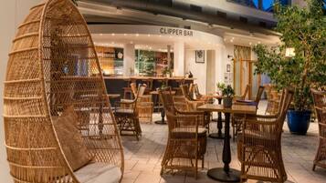 A cozy indoor seating area with wicker chairs and a hanging chair, a bar named 'CLIPPER BAR' in the background, and potted plants around.