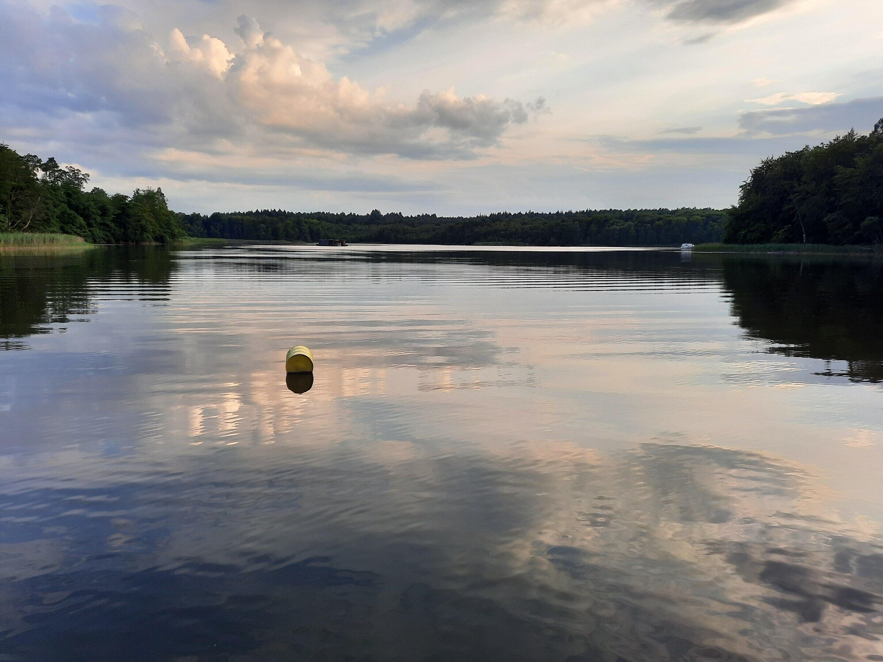Ein ruhiger See mit einem gelben Gegenstand, der auf dem Wasser schwimmt, umgeben von dichten Bäumen und einem bewölkten Himmel.