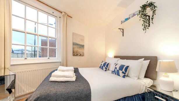 A bedroom with a large window, a bed with a blue skirt, a bedside table with a lamp, and a mirror. - Stylish 2 bed - Tower Hamlets (Greater London)