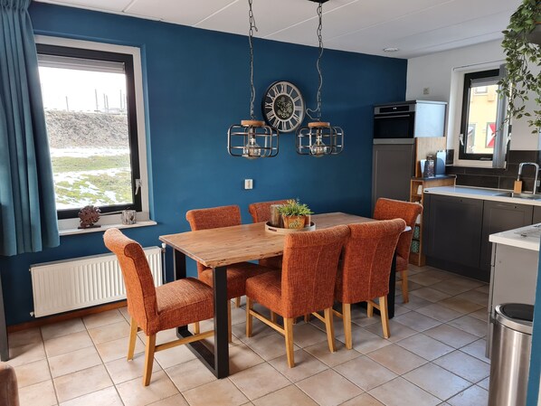 A dining area with a wooden table and orange chairs, a window with curtains, and a kitchen area with a refrigerator and sink.