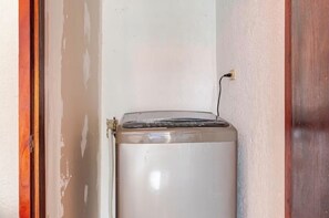 Een compacte, witte wasmachine in een hoek, met een houten deurkozijn en een stopcontact op de muur.
