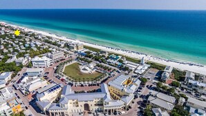 Luftaufnahme einer Küstenstadt mit einem zentralen Platz, Strand und verschiedenen Gebäuden, darunter ein großes Hotel mit einem kreisförmigen Innenhof.