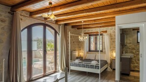 A bedroom with a wooden ceiling, a large window with curtains, a bed, a bedside table, and a view of the outdoors. - Countryside Villa with a Walk-in Free Form Pool (Rethymno)