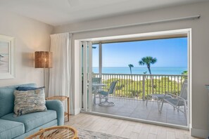 Ein Zimmer mit einer Couch, einem Tisch und Stühlen, einer großen Schiebeglasdoor zum Balkon mit Blick aufs Meer und Palmen.